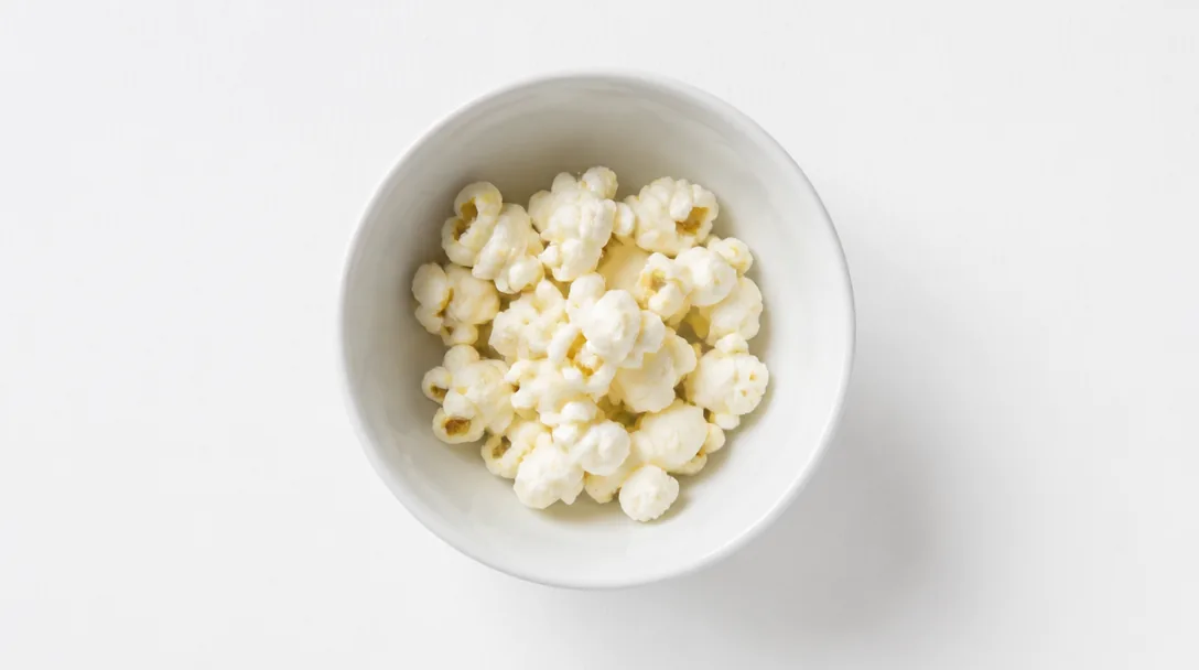 Liten portion luftpoppad popcorn i vit keramikskål på vit yta, ovanifrånperspektiv